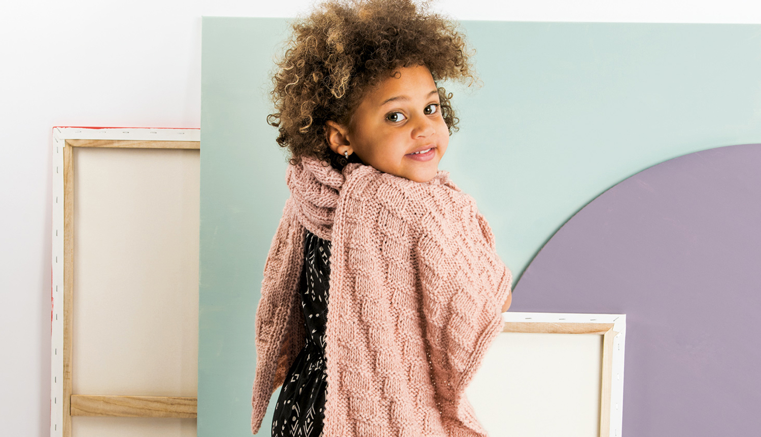 Strickanleitung Schal mit Strukturmuster: Kind trägt einen rosa Schal mit Zopfmuster vor pastellfarbenem Hintergrund.