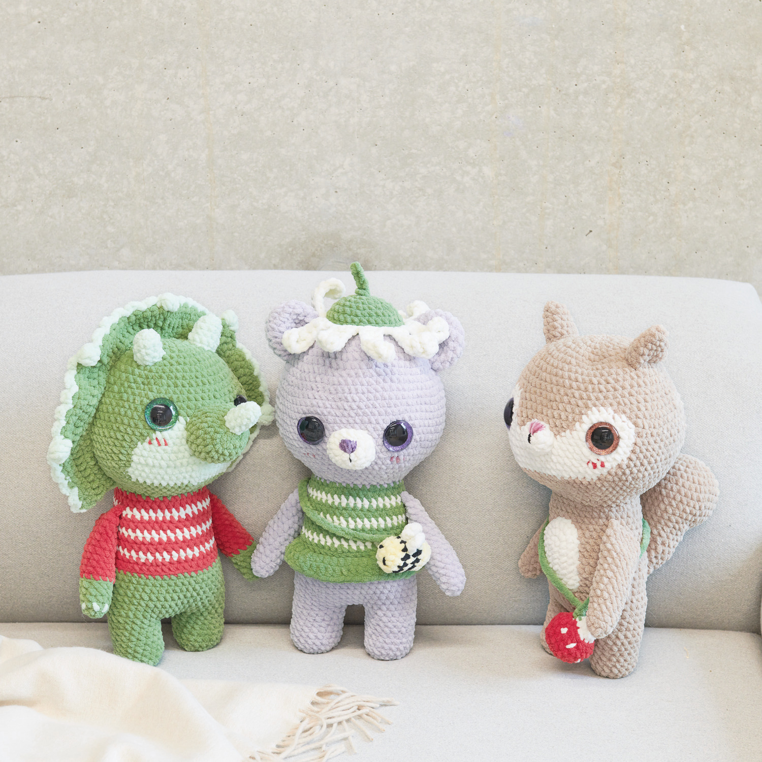 Häkelset Little Squirrel Modell 02: Drei gehäkelte Figuren, darunter ein Dinosaurier, ein Bär und ein Eichhörnchen, auf einem Sofa sitzend.