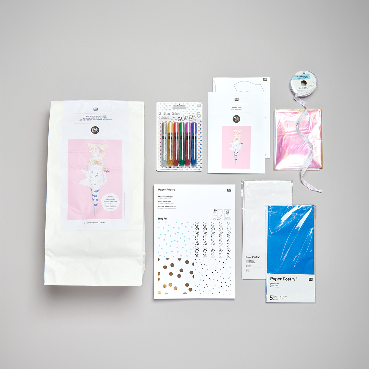 Ein Schultüten Bastelset, daneben ein Motivpapierblock, verpacktes Krepppapier, glitzer Stifte und eine Rolle Dekoband. Flatlay von Produkten.