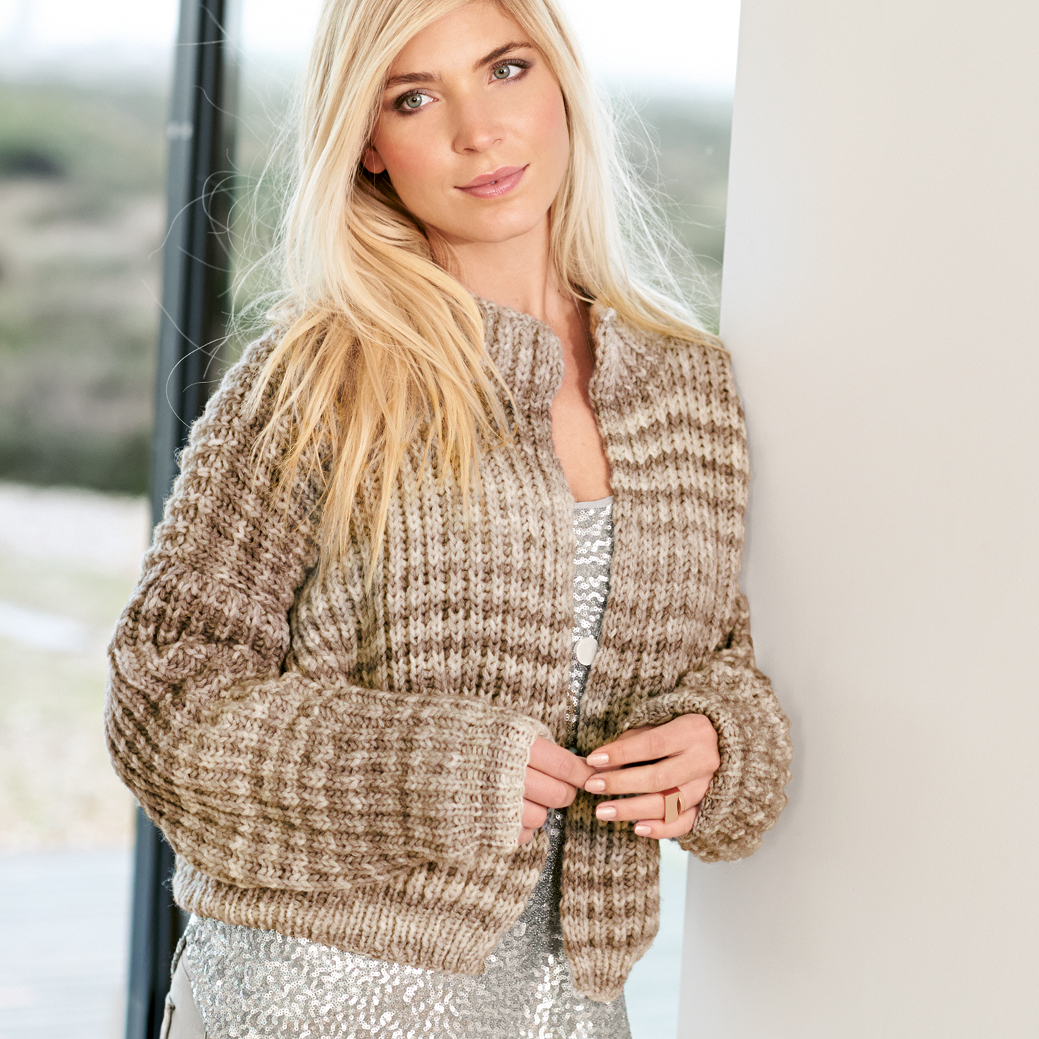 Strickanleitung Jacke aus Creative Light Melange: Frau trägt gestrickte Jacke in melierten Brauntönen, stehend vor einem Fenster.