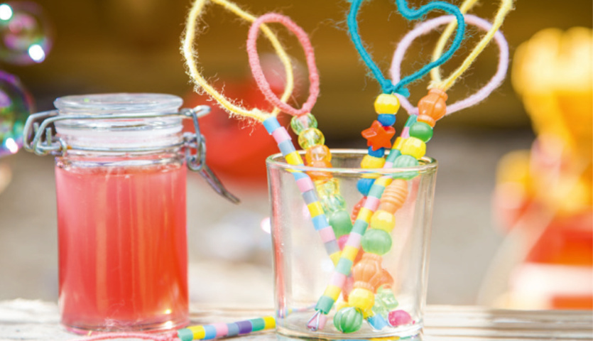 Bastelanleitung DIY-Seifenblasen: Bunte Stäbe mit Perlen und Draht in Glas, daneben rotes Seifenblasen-Gemisch im Glasbehälter.