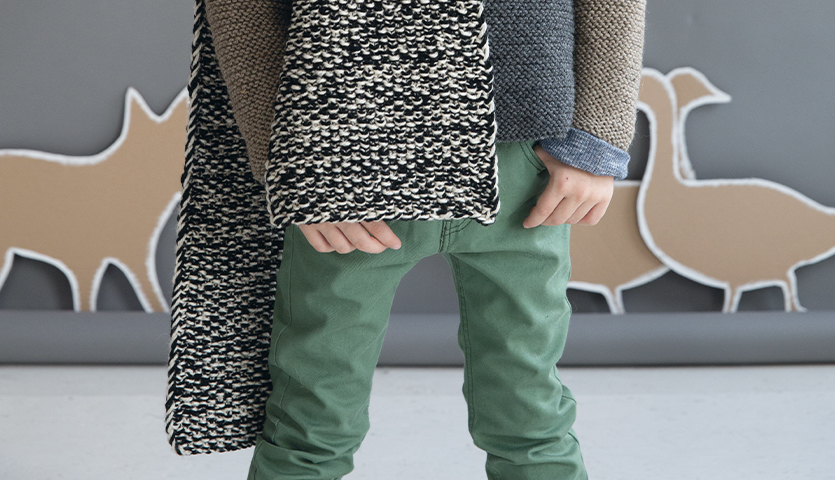 Strickanleitung Kinder-Perlmusterschal aus Soft Merino: Kind trägt schwarz-weißen Schal, kombiniert mit grüner Hose und grauem Pullover.