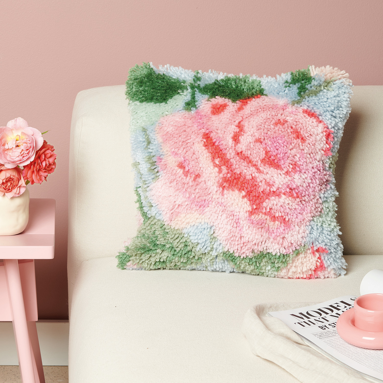 Rico Hooki Hooki Knüpfset Kissen Rose zeigt ein flauschiges Kissen mit Rosenmotiv auf einem Sofa.
