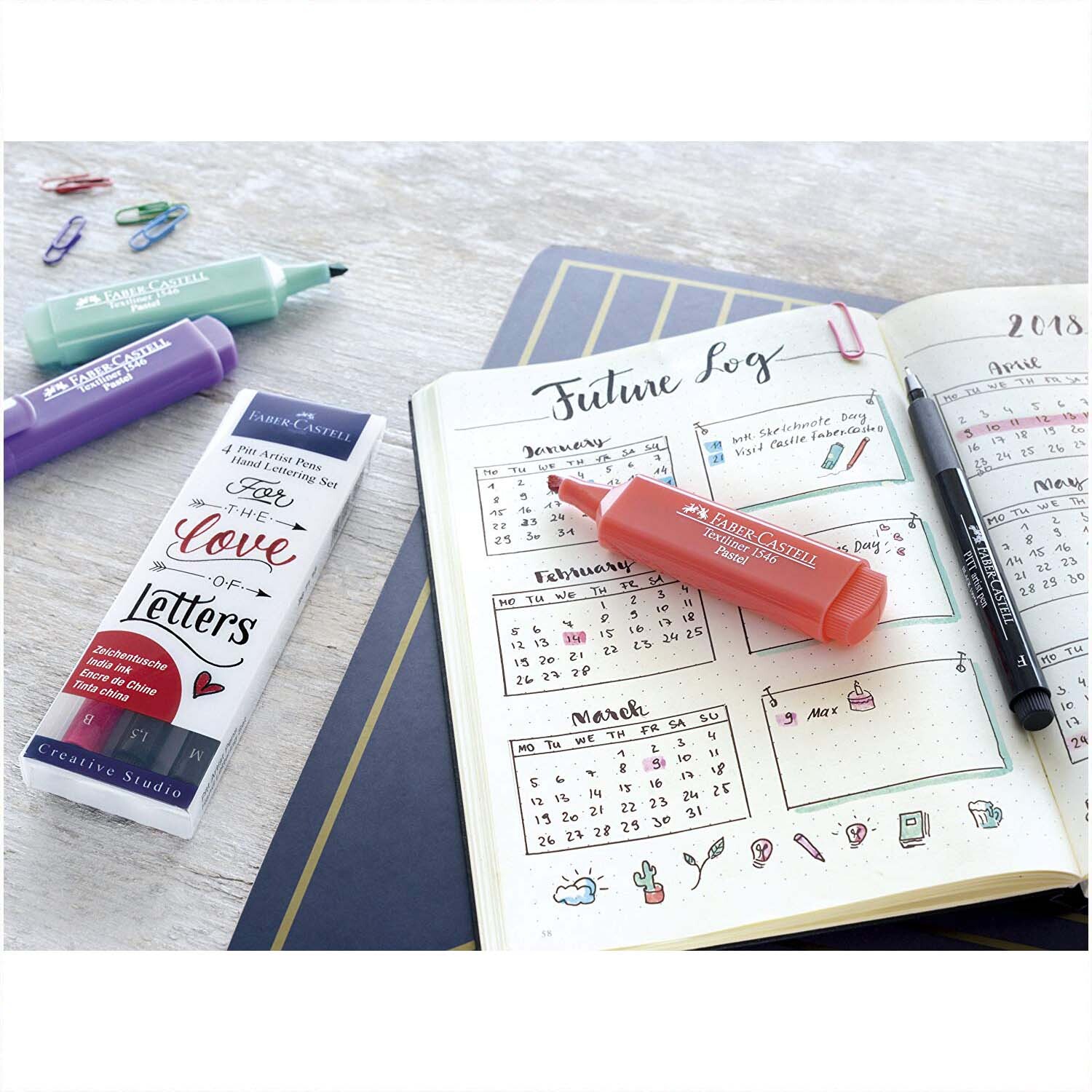 Tuschestift Pitt Artist Pen Handlettering 4 Stück von Faber Castell neben einem geöffneten Planer, umgeben von bunten Stiften und Büroklammern.