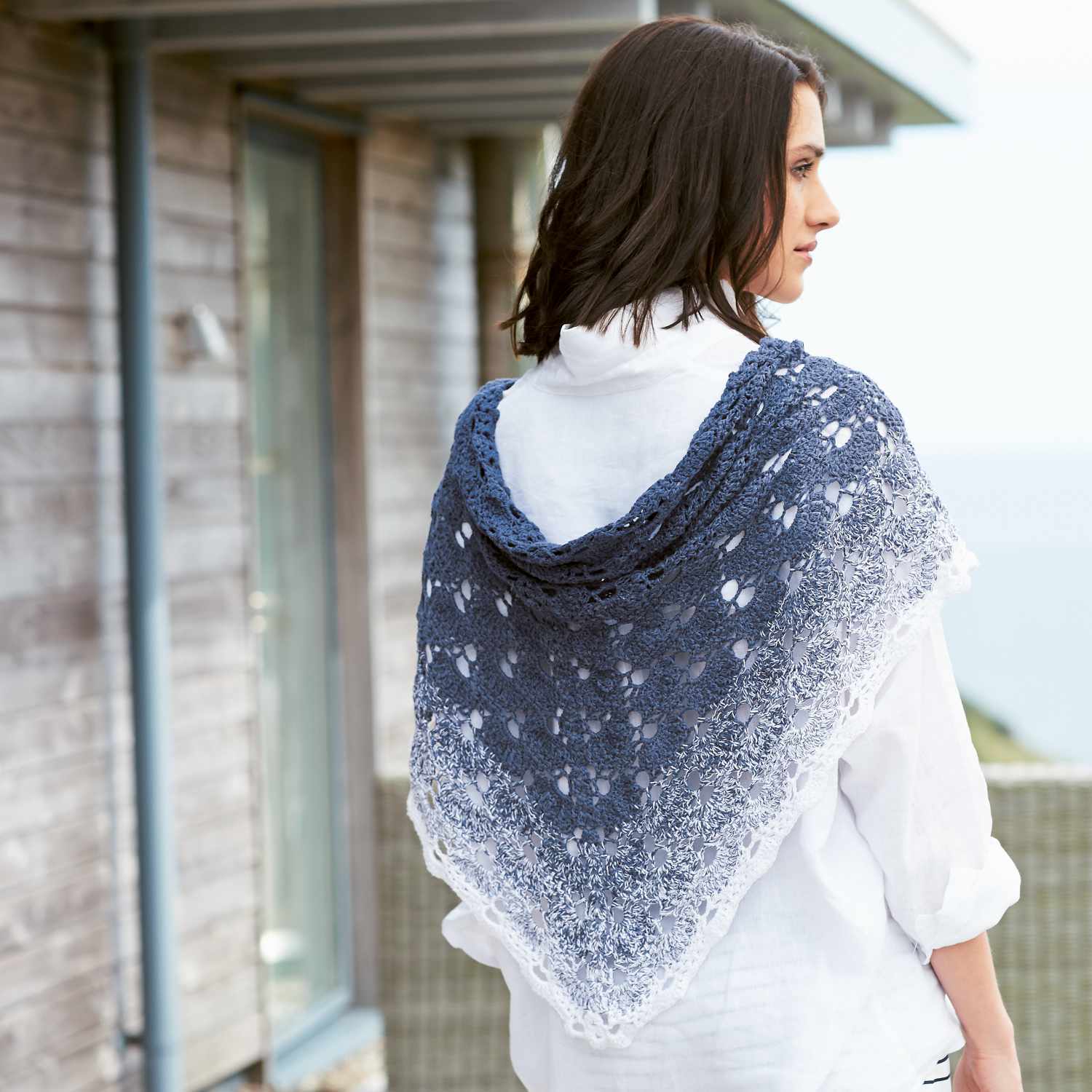 Häkelanleitung Dreieckstuch aus Creative Cotton Dégradé: Frau trägt blau-weißes Tuch auf Terrasse.