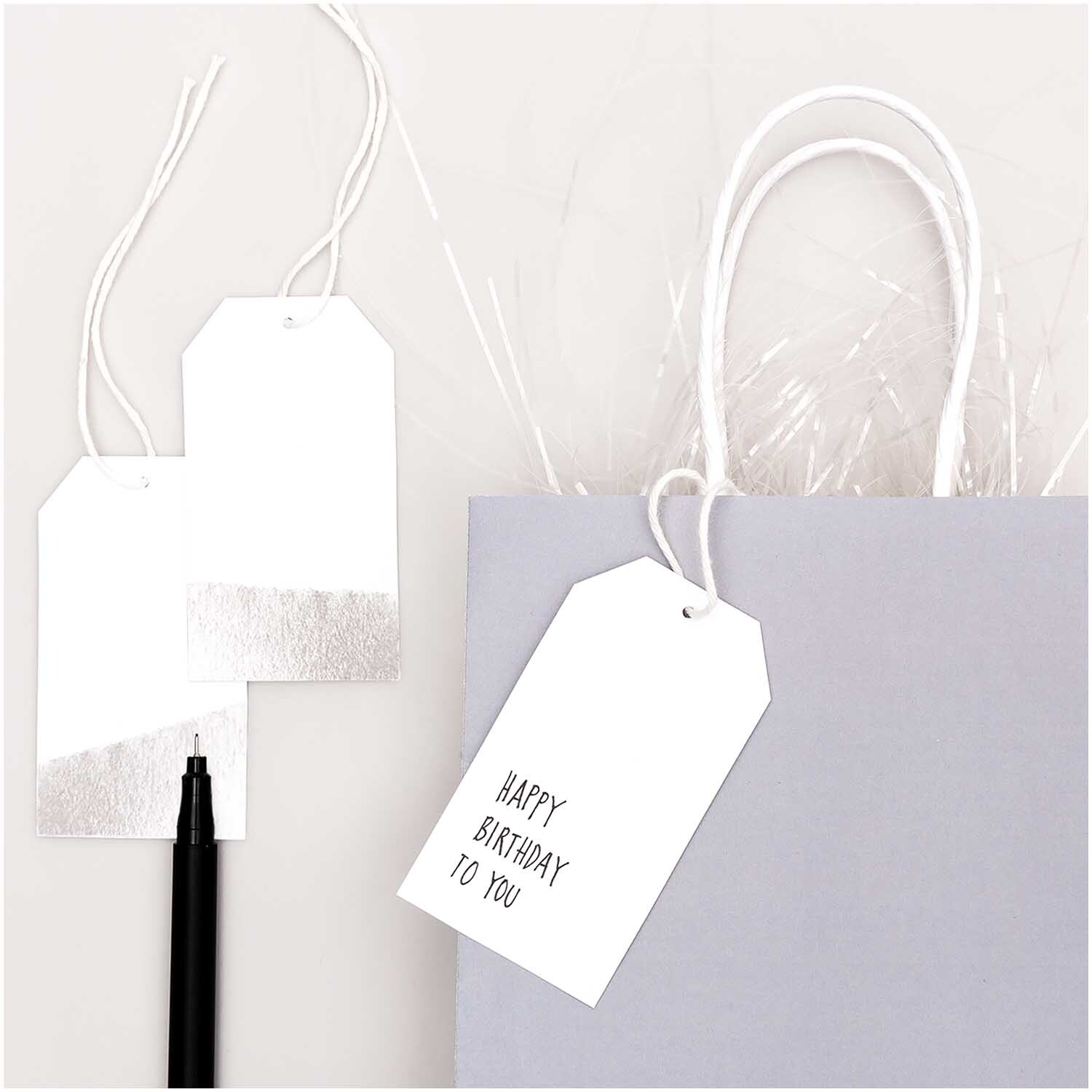 Paper Poetry Papieranhänger in Weiß-Silber von Rico Design, dekorativ an einer Geschenktüte befestigt. Ideal für Geburtstagsgeschenke.