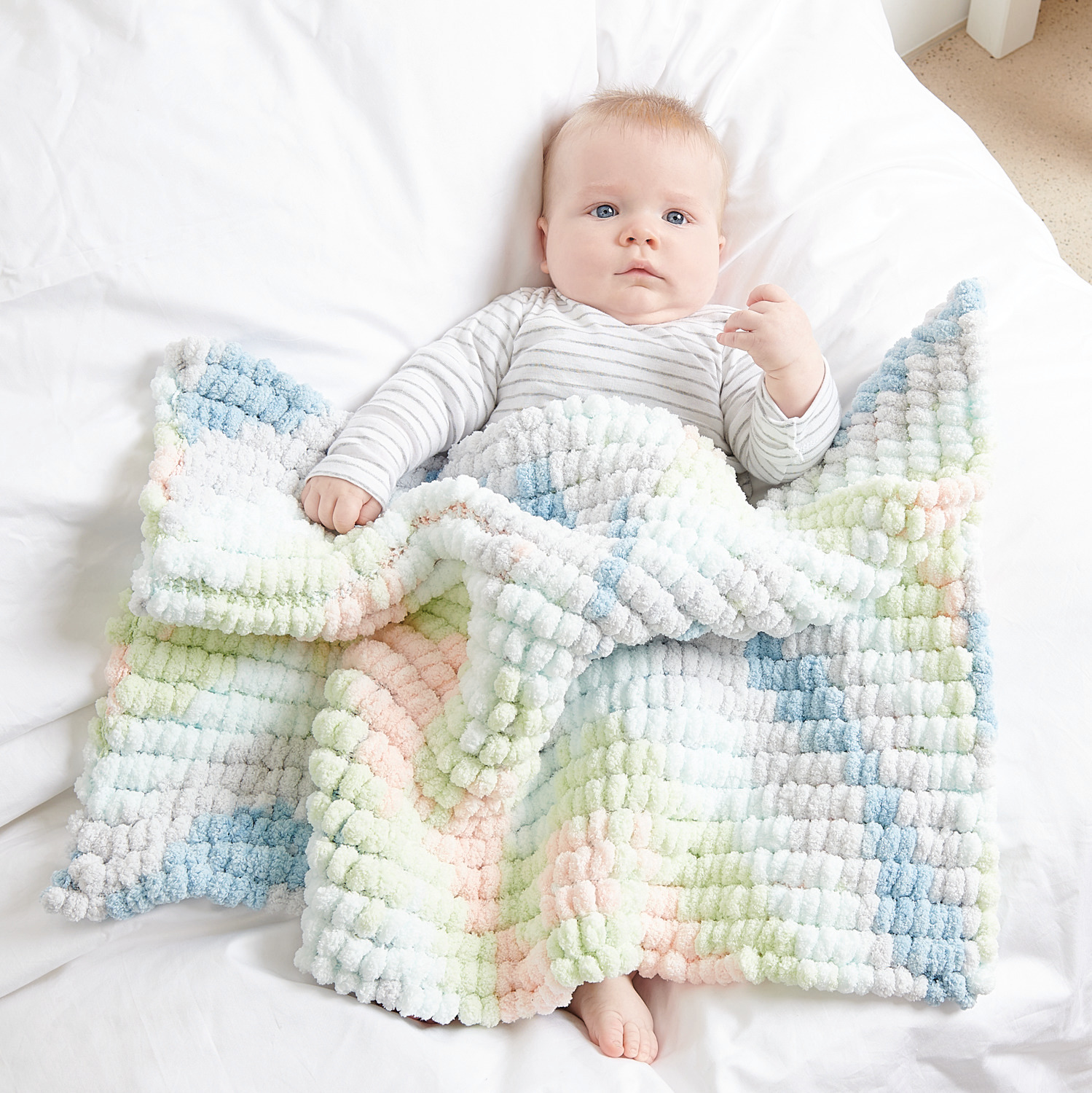 Creative Pompon Print: Ein Baby liegt auf einer weißen Decke mit einer pastellfarbenen, strukturierten Strickdecke von Rico Design.