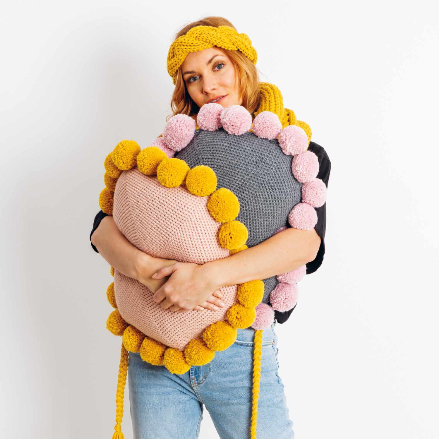 Häkelanleitung Kissen aus Essentials Mega Wool chunky: Frau hält buntes Kissen mit Pompons in Rosa, Grau und Gelb.