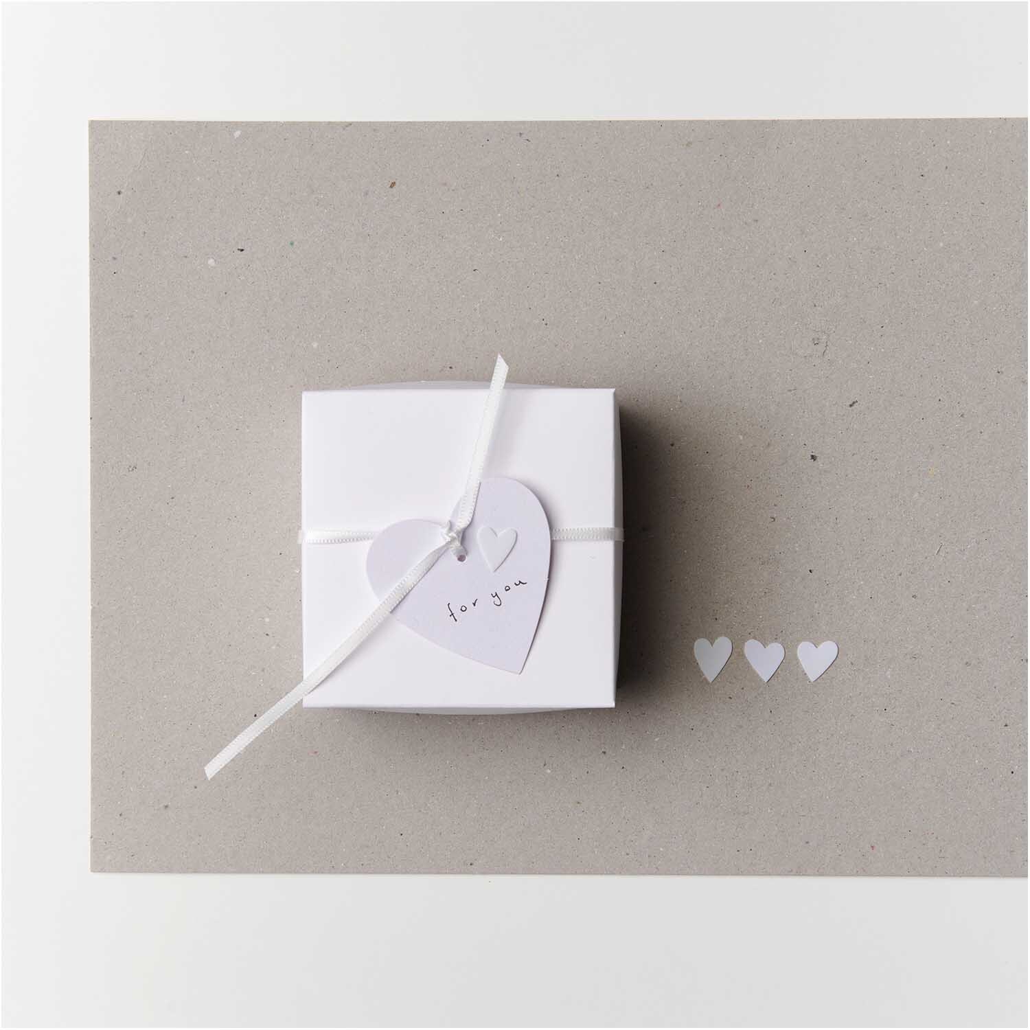 Flitter Herz 100 Stück: Weiße Papierherzen von Rico Design neben einer Geschenkbox auf grauem Hintergrund.