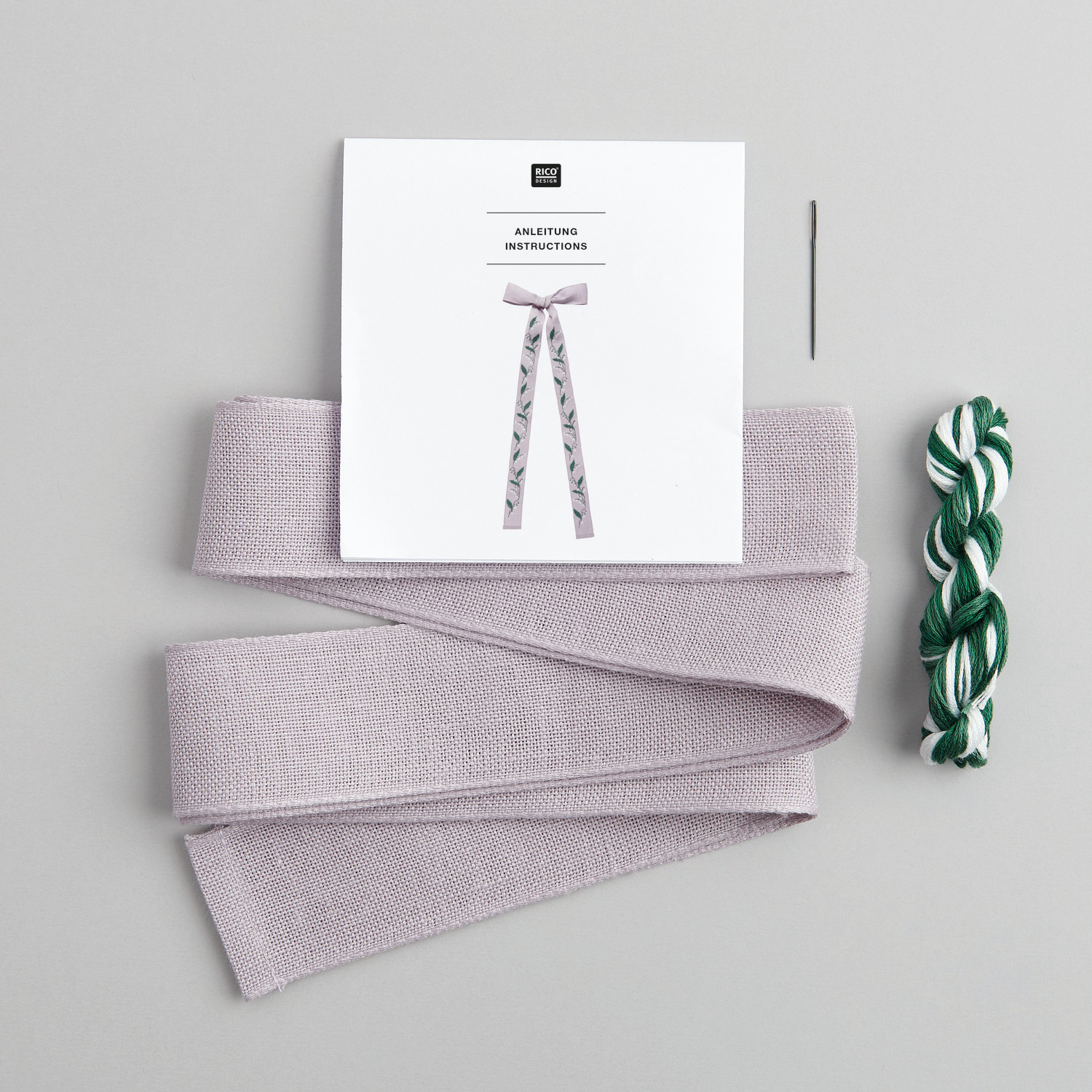 Stickpackung Schleife Maiglöckchenranke von Rico Design mit Anleitung, Nadel und Garn in Grün und Weiß auf grauem Hintergrund.