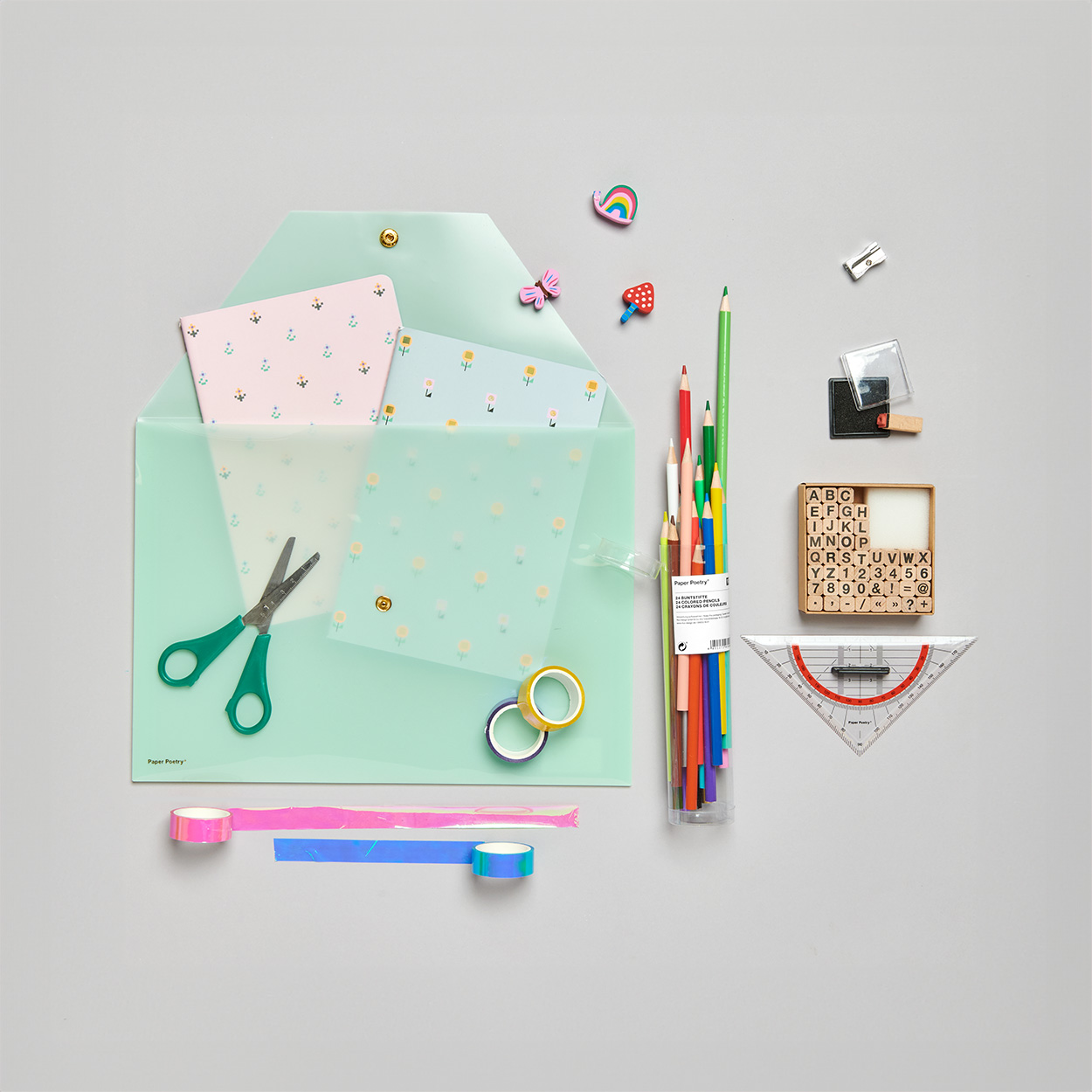 Grüne Mappe mit Motivpapier gefüllt, darauf eine Schere und Tapes und daneben Buntstifte, Stempel, ein Geodreieck und Radiergummis. Flatlay von Produkten.