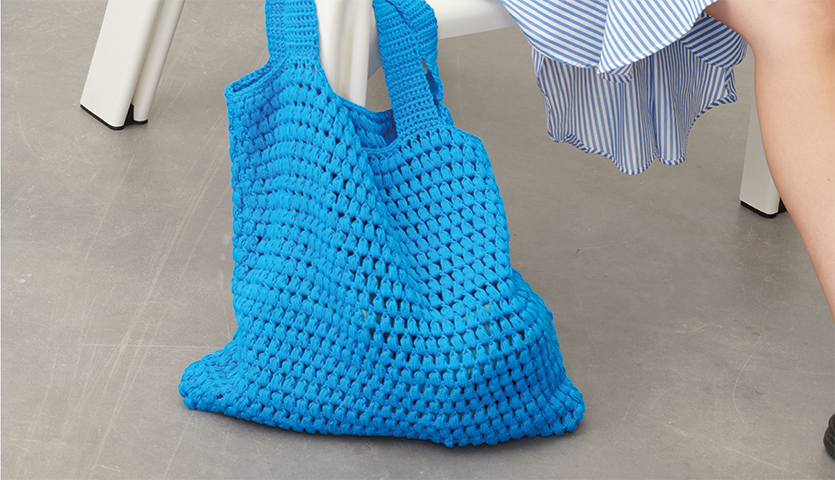 Häkelanleitung Tasche aus Creative Cotton aran: Eine blaue, gehäkelte Tasche auf grauem Boden.