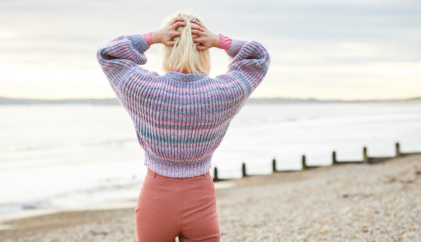 Strickanleitung Pullover aus Creative Melange Chunky: Frau am Strand in pastellfarbenem Pullover, der Rückenansicht zeigt.