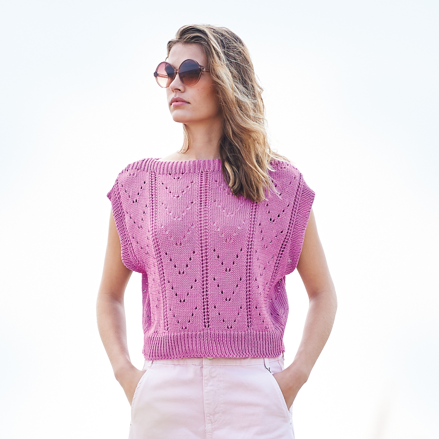 Strickanleitung Lochmuster-Top: Frau trägt ein rosa Lochmuster-Top aus Baumwolle, kombiniert mit Sonnenbrille und heller Hose.