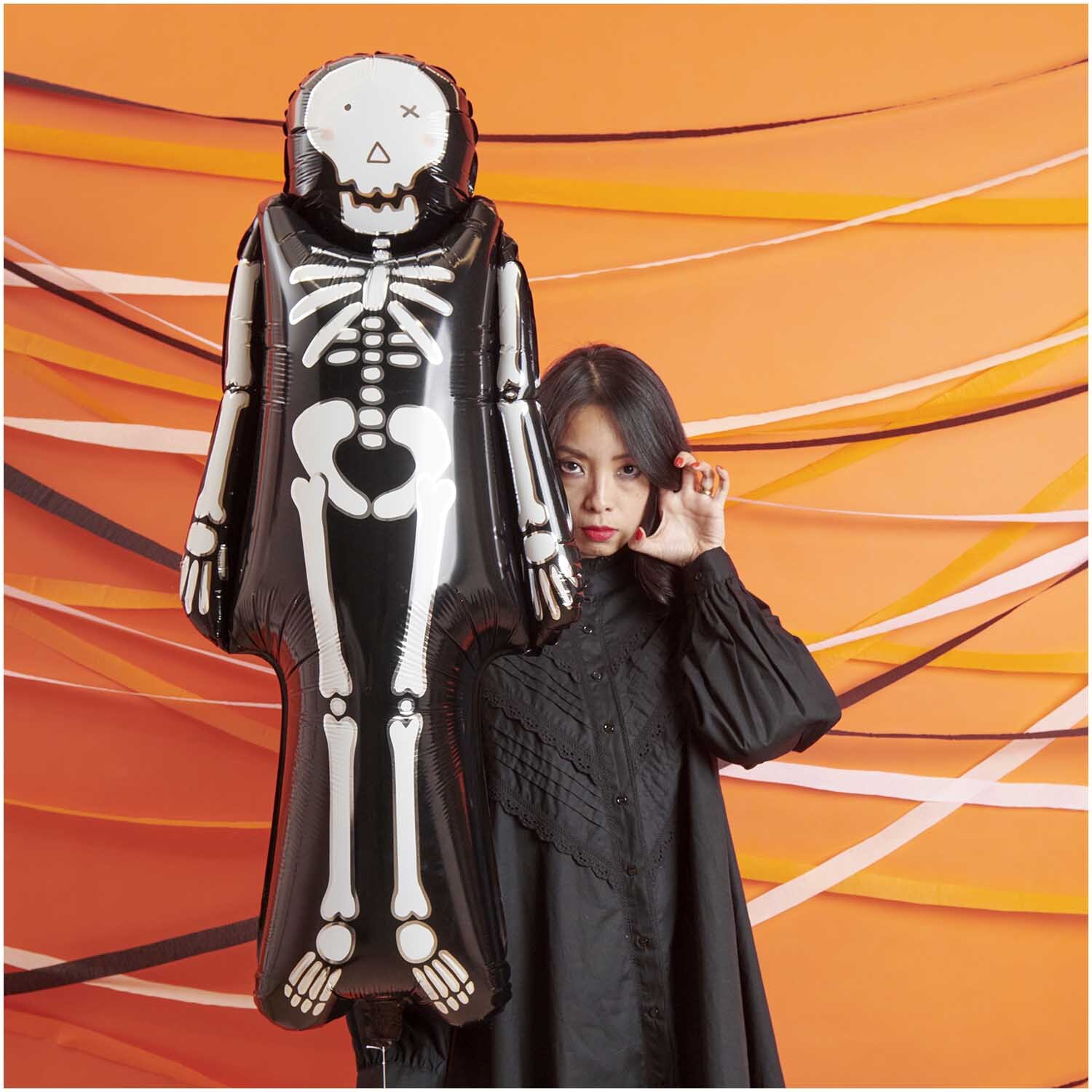 Folienballon Skelett 110x48cm von Rico Design vor orangefarbenem Hintergrund mit Frau in Schwarz.