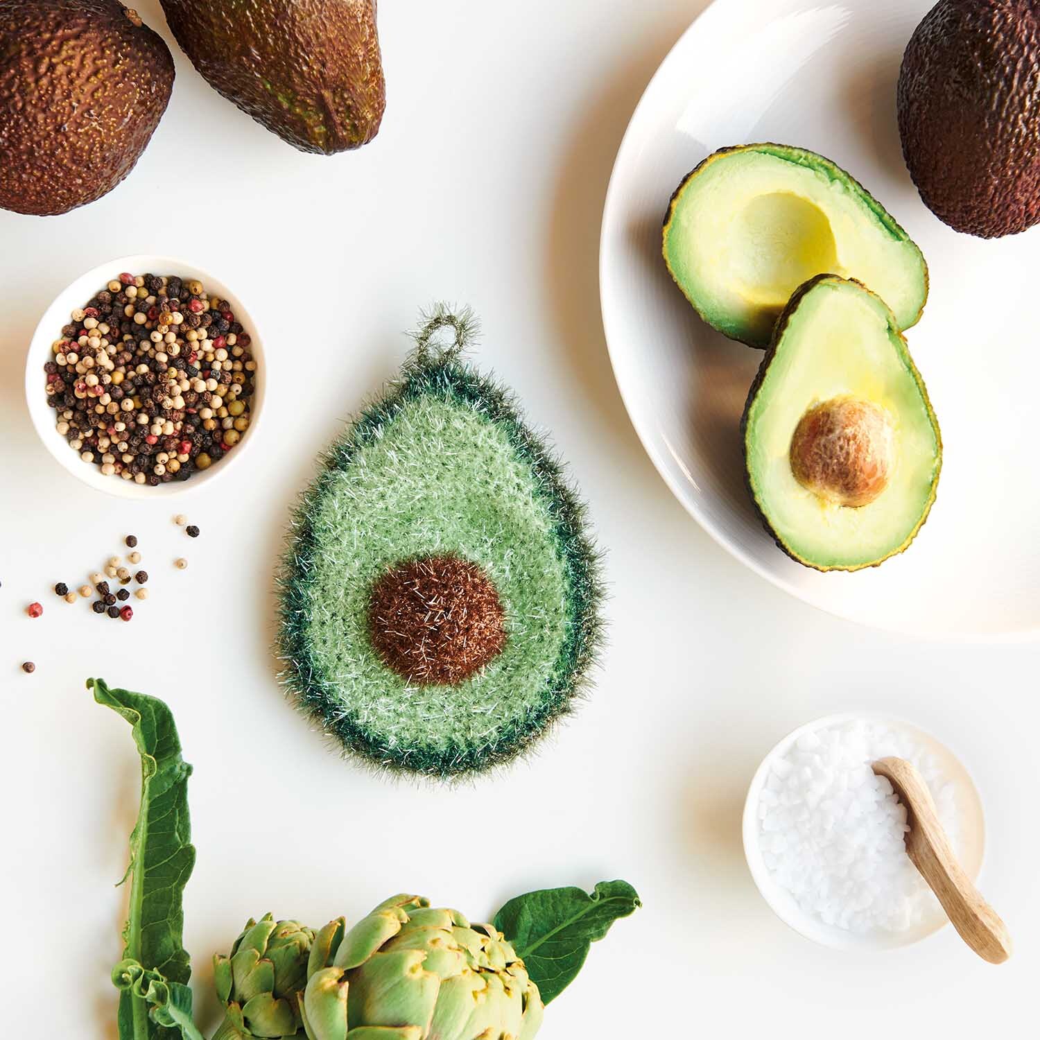 Häkelanleitung Avocado aus Creative Bubble: Grüne Avocado-Häkelarbeit neben frischen Zutaten auf weißem Hintergrund.