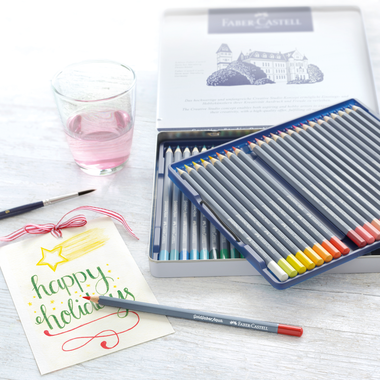 Goldfaber Aqua Aquarellstifte im Metalletui, 48-teilig, mit farbenfrohen Stiften und einem Glas Wasser auf einem Tisch.