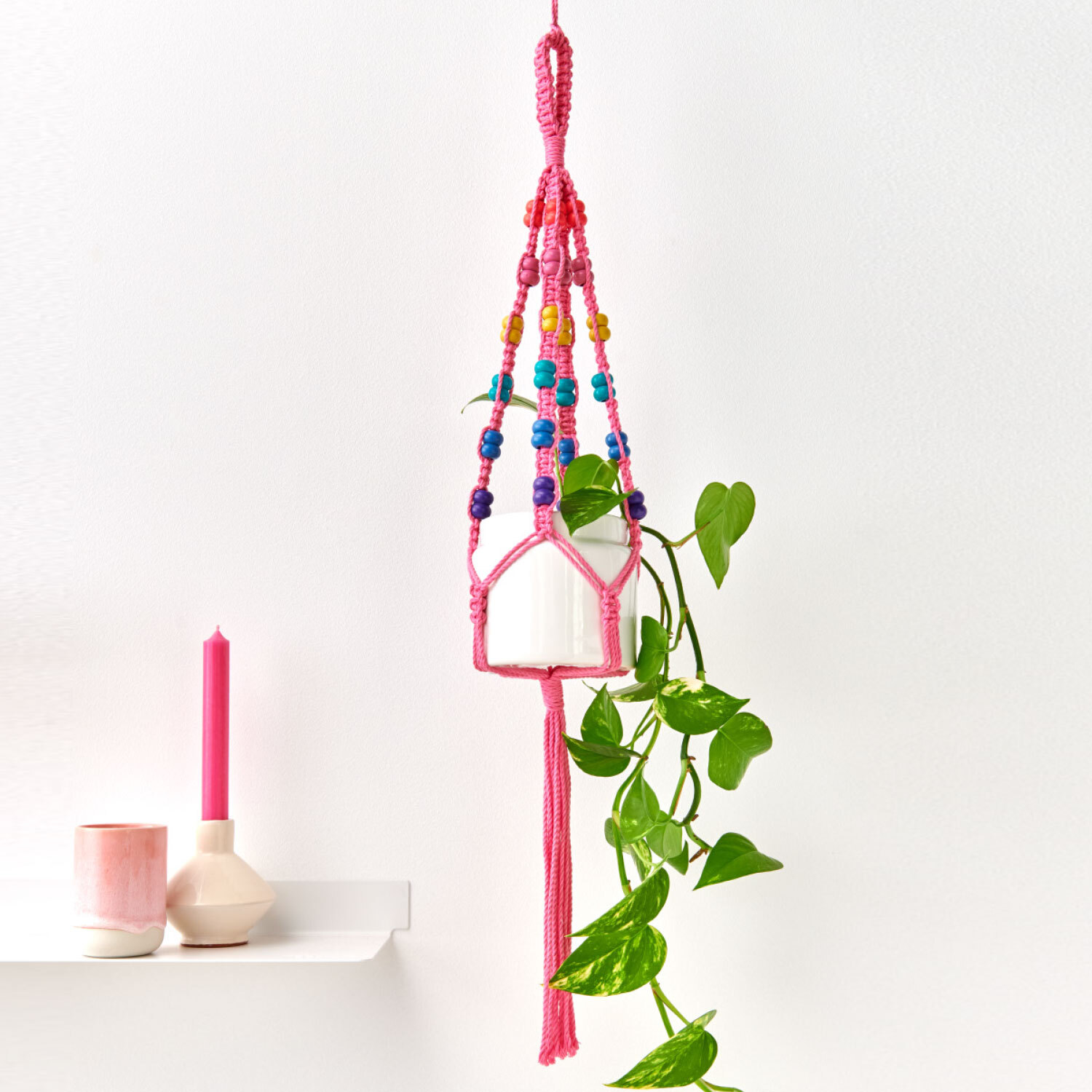 Anleitung Makramee-Blumenampel mit bunten Holzperlen, hängend an einer weißen Wand mit Pflanze.