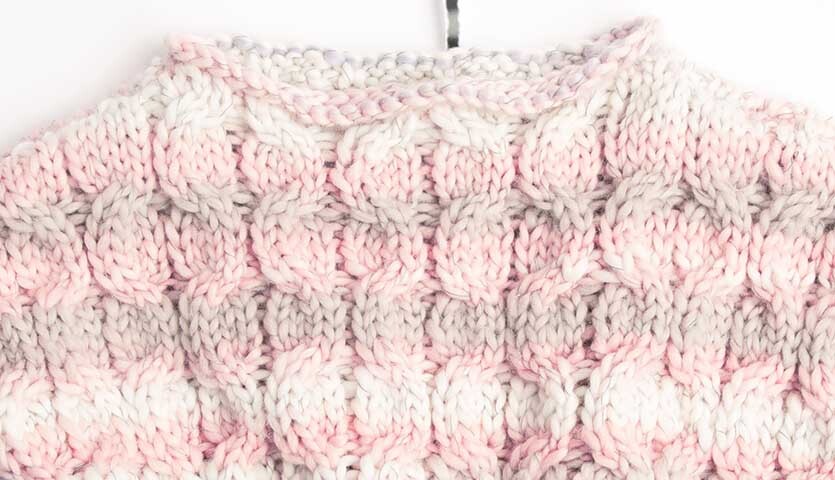 Strickanleitung Poncho aus Creative Glühwürmchen Print: Detailaufnahme eines gestrickten Ponchos in Rosa und Grau mit Zopfmuster.