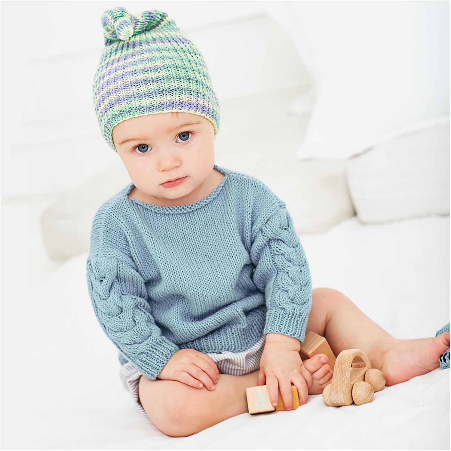 Rico Baby Cotton Soft dk: Kleinkind in gestricktem Pullover und Mütze, spielt mit Holzspielzeug.