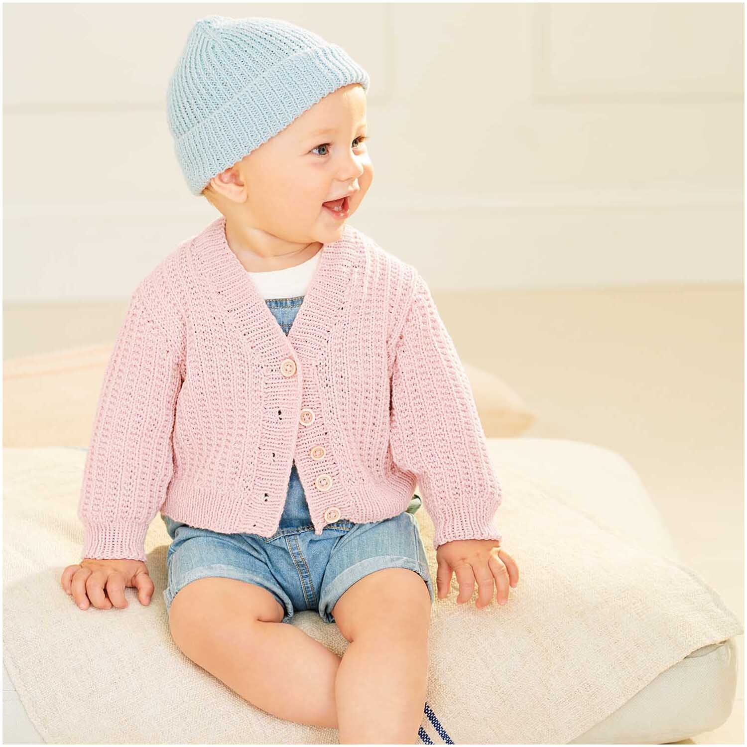 Baby Organic Cotton: Ein lächelndes Baby trägt eine rosa Strickjacke von Rico Design und eine blaue Mütze, sitzt auf einem Kissen.