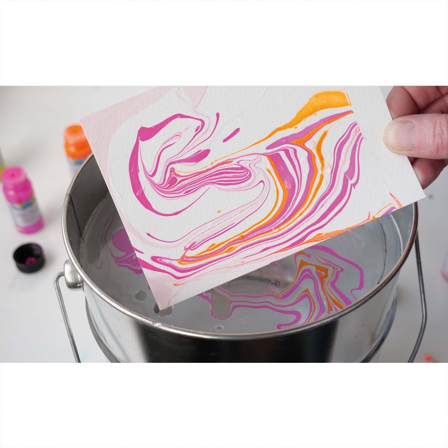 Magic Marble Marmorierfarben Set Love Neon 6x20ml zeigt eine Hand, die ein marmoriertes Papier in leuchtenden Neonfarben aus einem Wasserbad hebt.