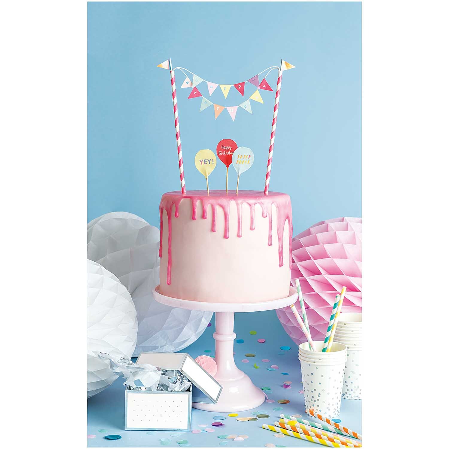 Kuchendekoration Happy Birthday pastell: Ein rosa Drip Cake mit bunten Wimpeln und Ballons auf einem Tisch mit Partyzubehör.