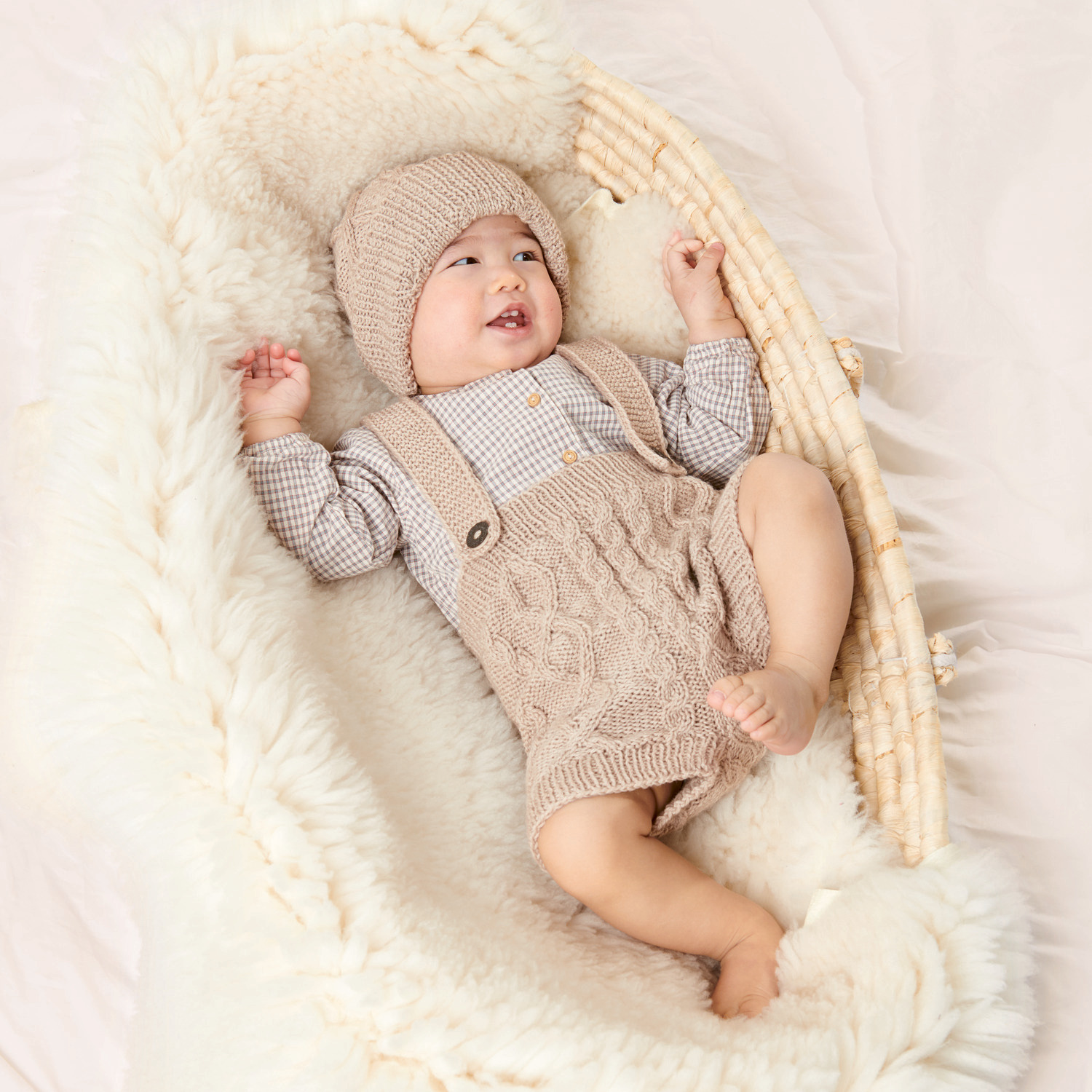 Strickset Babymütze Modell 14: Ein Baby trägt eine gestrickte Mütze und Latzhose in einem weichen, beigen Farbton, liegend in einem kuscheligen Korb.