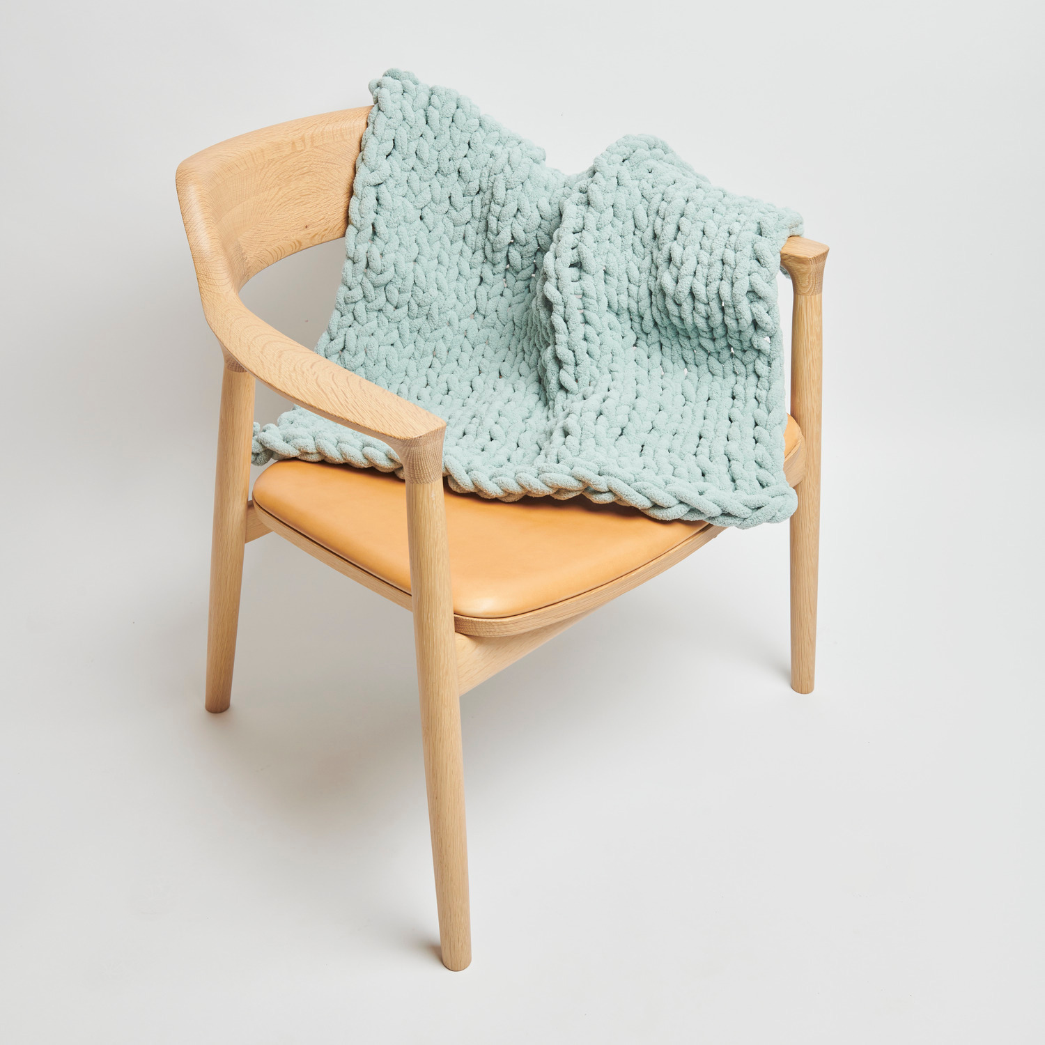 Chenillove Super Big: Weiche, salbeifarbene Strickdecke aus Chenille auf einem Holzstuhl drapiert.