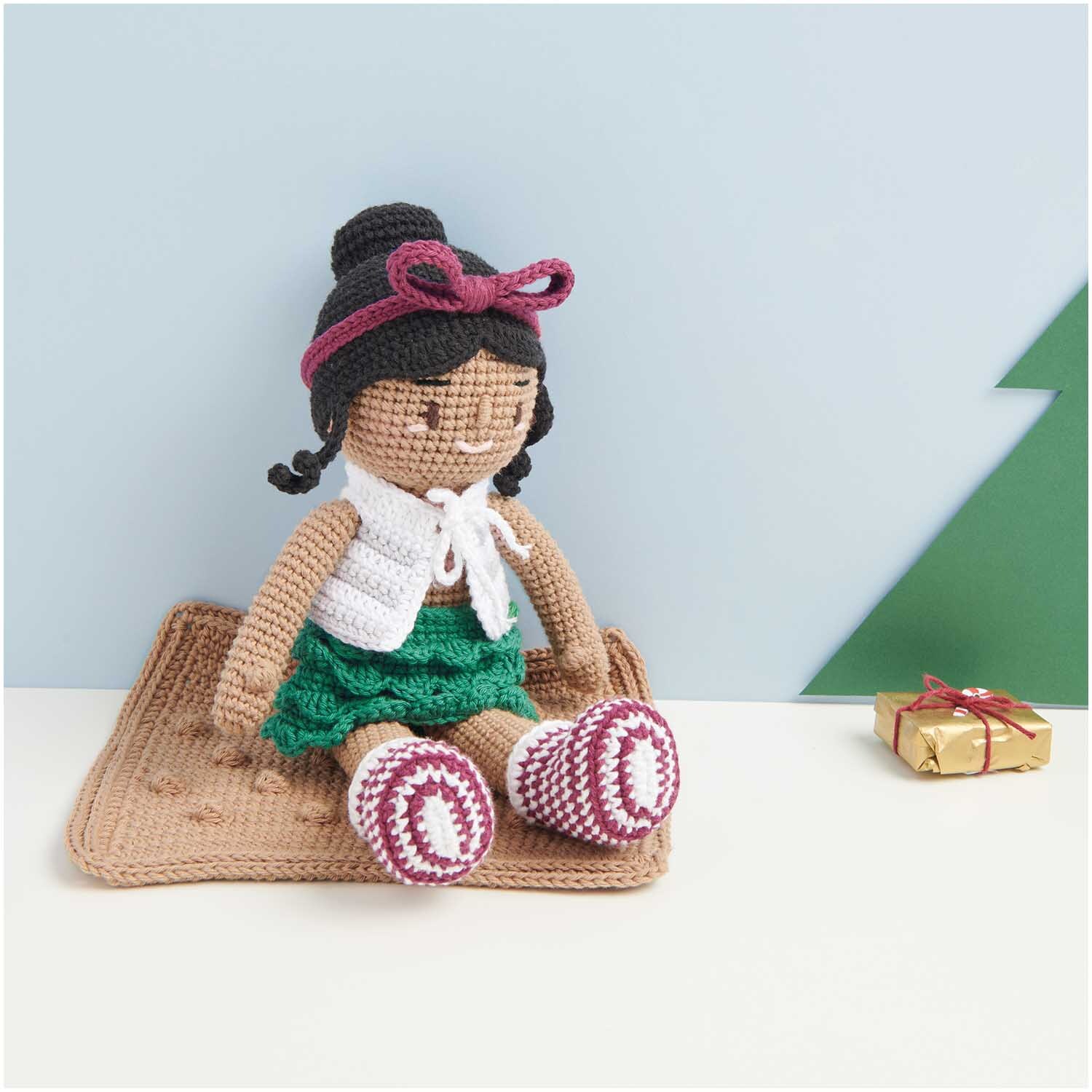 Creative Ricorumi dk: Gehäkelte Puppe mit bordeauxfarbenem Stirnband, sitzt auf einer Decke neben einem Geschenk.