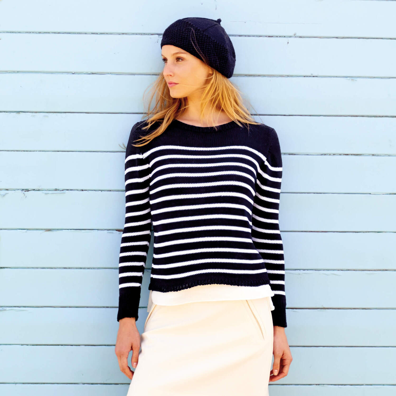 Strickanleitung Pullover und Baskenmütze im Navy-Look: Frau in gestreiftem Pullover und passender Mütze vor hellblauer Wand.