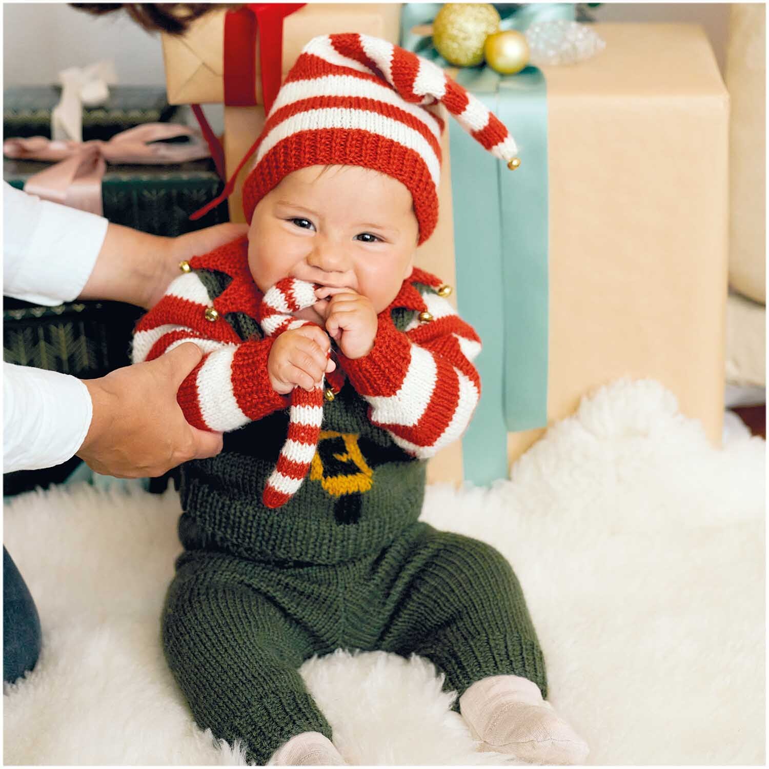 Essentials Merino Plus dk: Baby in gestreiftem Strickanzug mit Mütze, umgeben von Geschenken, auf weichem Teppich.