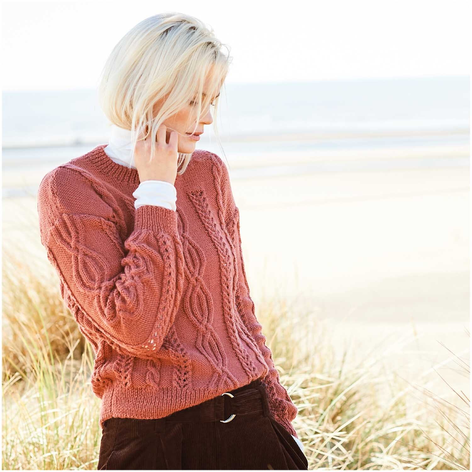 Strickanleitung Pullover aus Essentials Merino Plus dk: Frau am Strand in einem gestrickten Pullover mit Zopfmuster, ideal für kühle Tage.