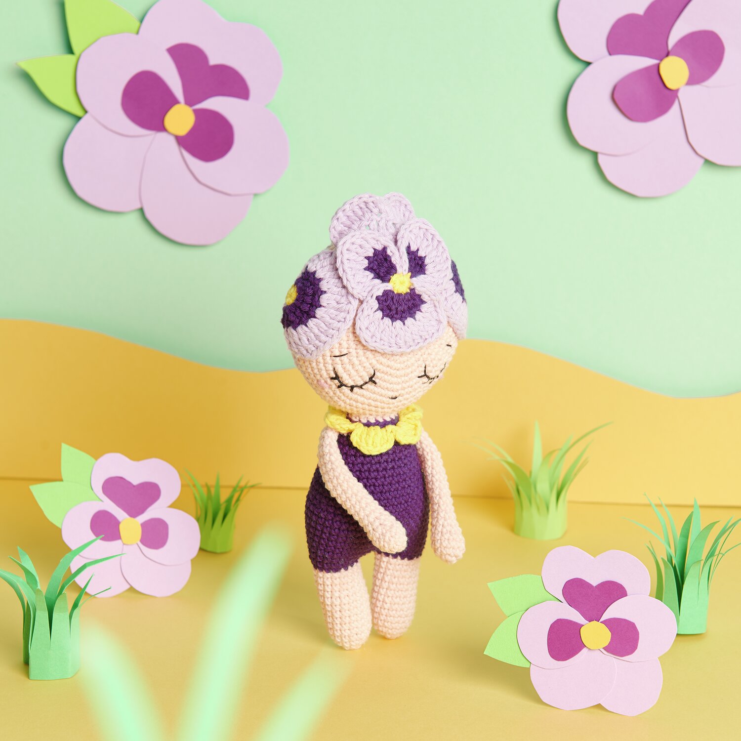 Creative Ricorumi dk Häkelpuppe in Lila und Flieder, umgeben von Papierblumen auf gelbem Boden.