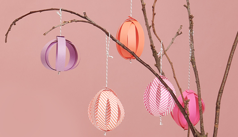 Bastelanleitung Ostereier aus Fröbelstreifen: Bunte Papierostereier hängen an einem Ast vor rosa Hintergrund.
