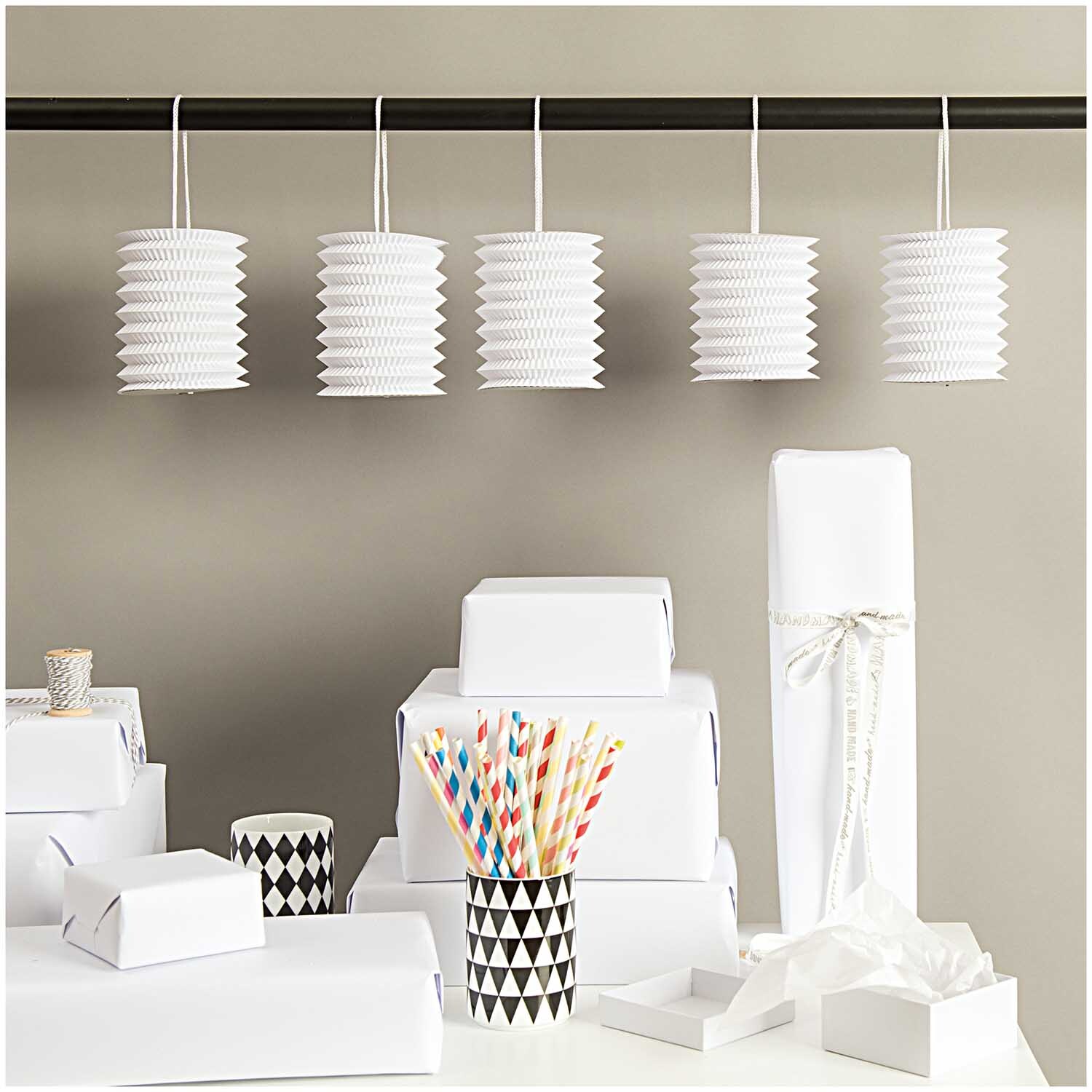 Zuglaterne weiß 10x13cm Papier 5 Stück von Rico Design, dekorativ hängend über einem Tisch mit Geschenkverpackungen und bunten Strohhalmen.
