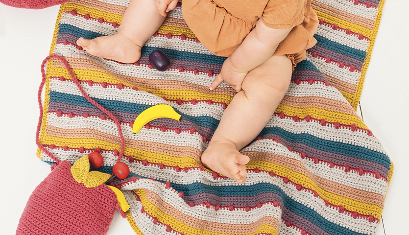 Häkelanleitung Decke aus Baby Cotton Soft DK: Bunte, gestreifte Decke mit Baby und Spielzeug.