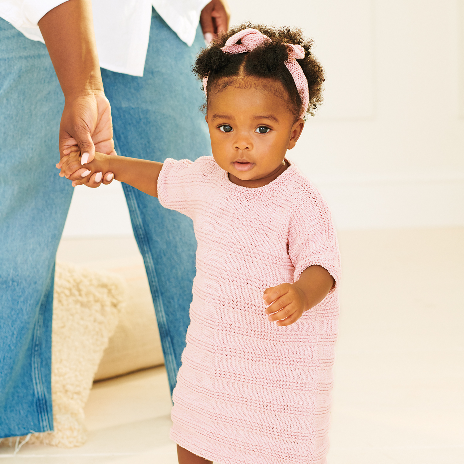 Strickanleitung Kleid und Stirnband aus Baby Organic Cotton: Ein Kleinkind trägt ein rosa gestricktes Kleid und Stirnband, gehalten von einem Erwachsenen.