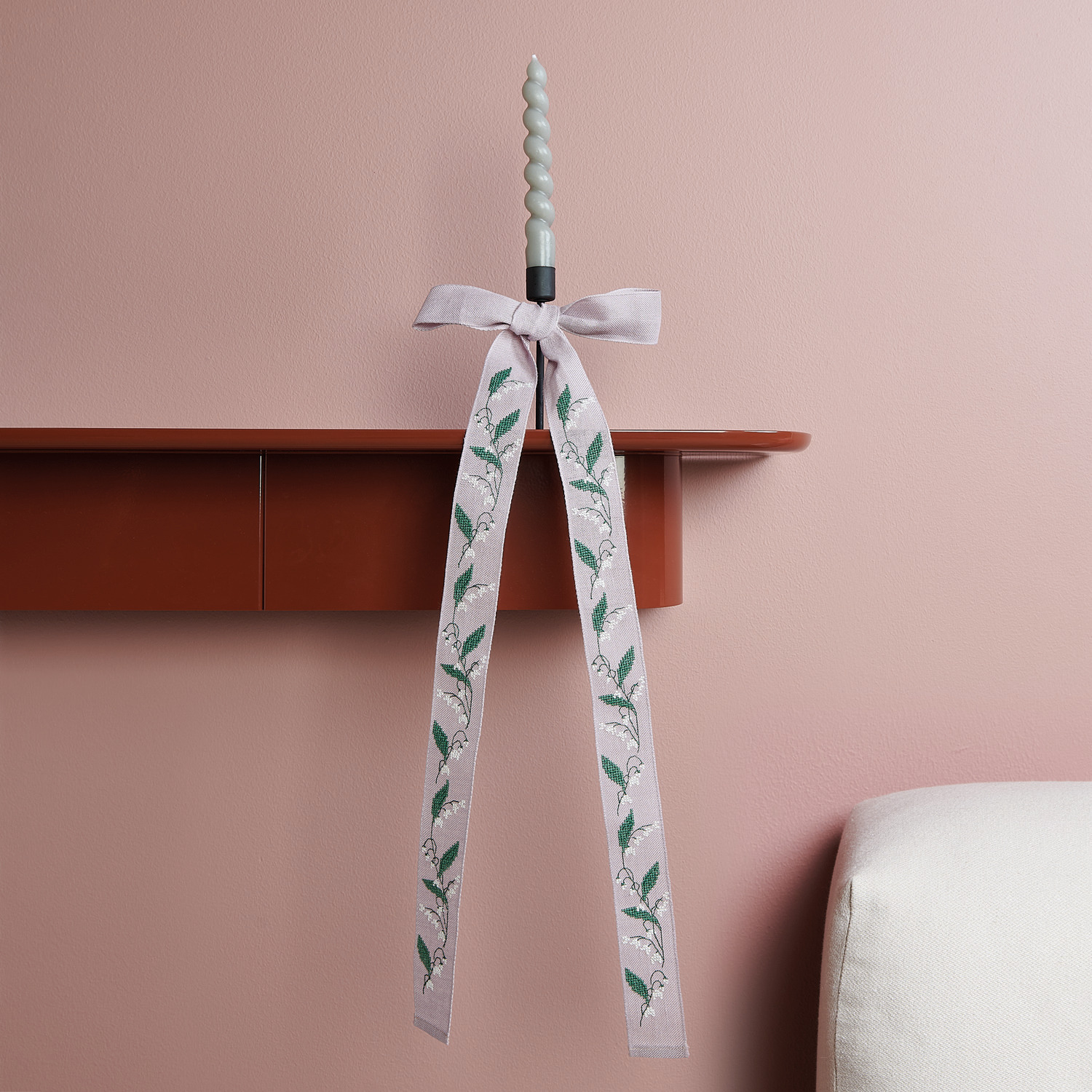 Stickpackung Schleife Maiglöckchenranke von Rico Design: Elegante Schleife mit Maiglöckchenmotiv an einer Kerze auf einem Regal.
