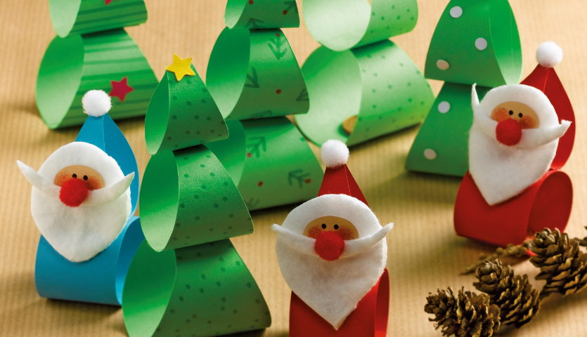 Bastelanleitung Xmas-Wackelfiguren: Bunte Papierfiguren von Weihnachtsmännern und Tannenbäumen auf einem Tisch.