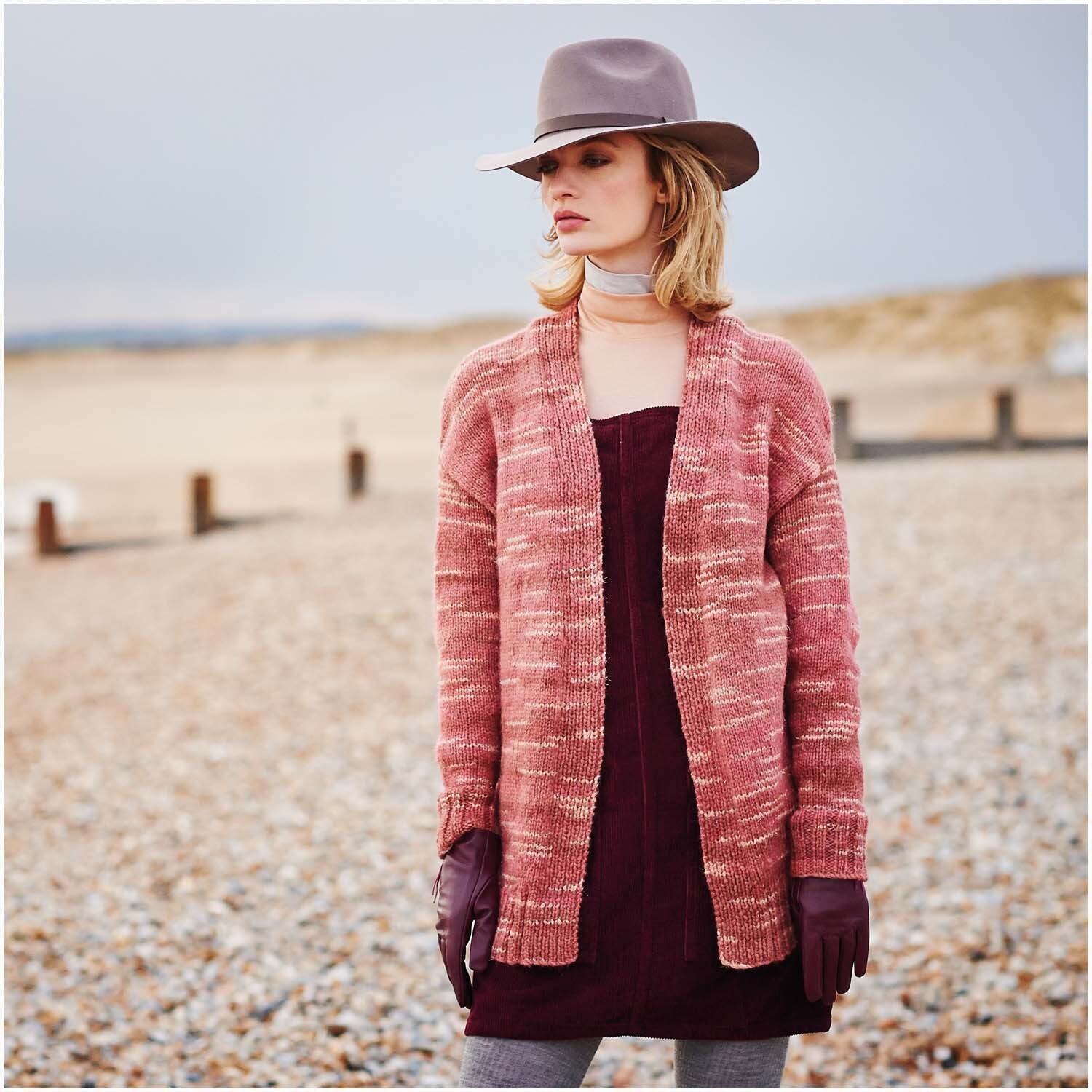 Creative Light Melange Glitz: Frau am Strand in langem Strickcardigan, kombiniert mit Hut und Handschuhen.