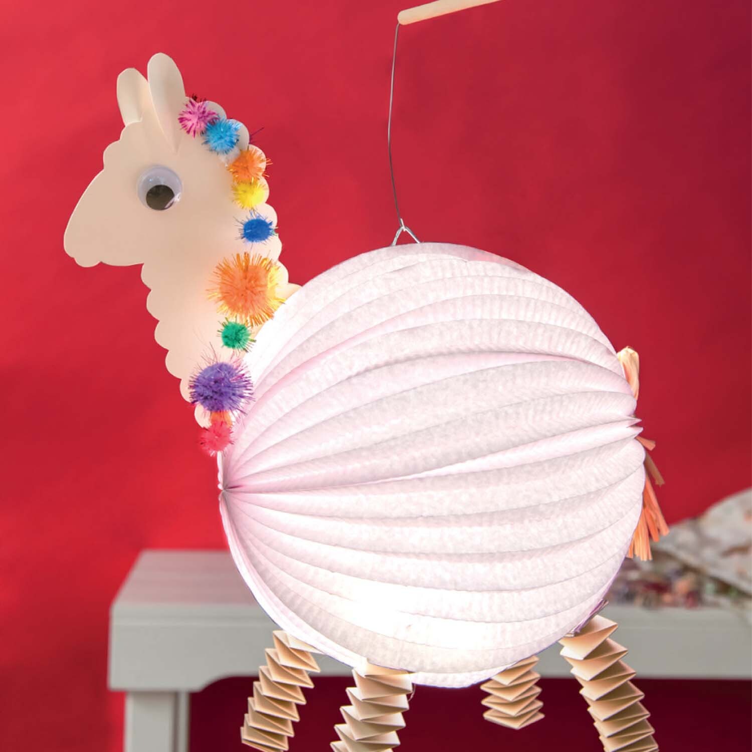 Bastelanleitung Lama-Laterne: Eine kreative Laterne in Lama-Form mit bunten Pompons und Wellpappe-Beinen vor rotem Hintergrund.
