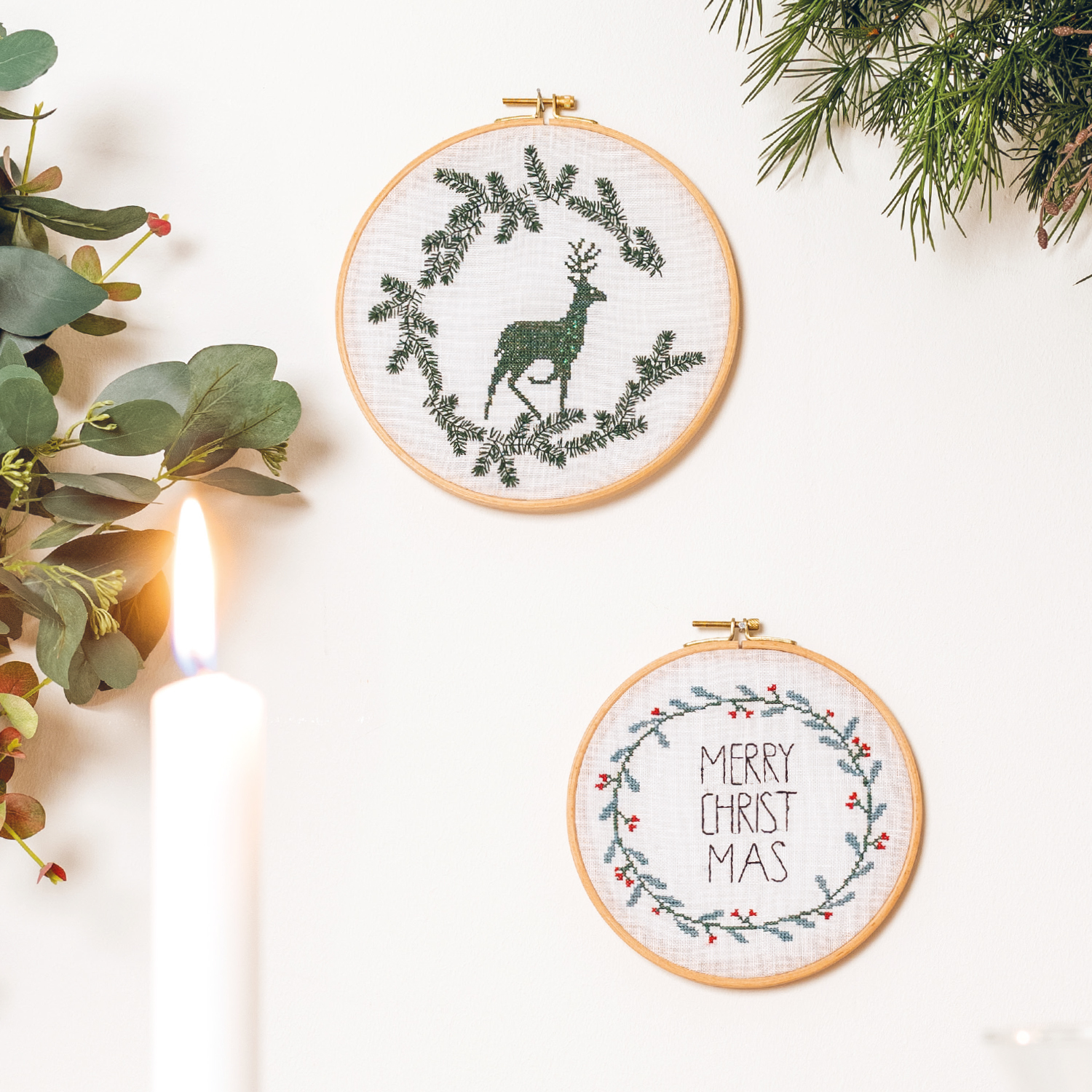 Stickanleitung Weihnachtliche Homedeko im Stickring: Zwei bestickte Ringe mit Hirschmotiv und Weihnachtsgruß, umgeben von grünen Zweigen und Kerze.