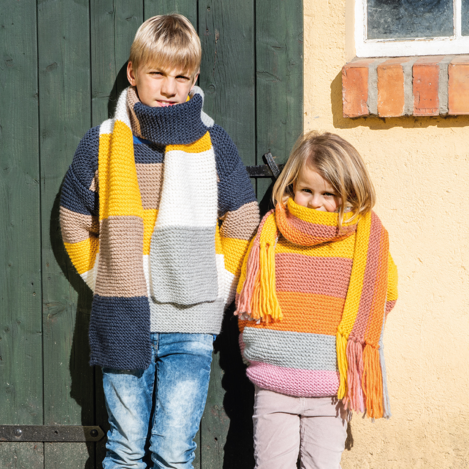 Strickanleitung Schal und Pullover aus Essentials Super chunky: Zwei Kinder tragen bunte, gestrickte Schals und Pullover vor einer Holztür.