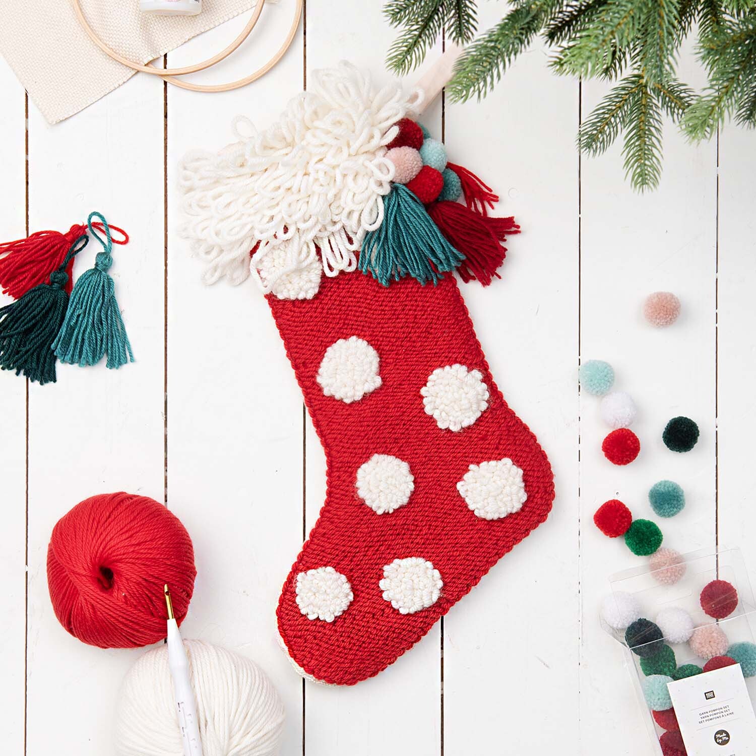 Anleitung gepunkteter Punch Needle Weihnachtsstrumpf auf weißem Hintergrund mit Garn und Pompons.