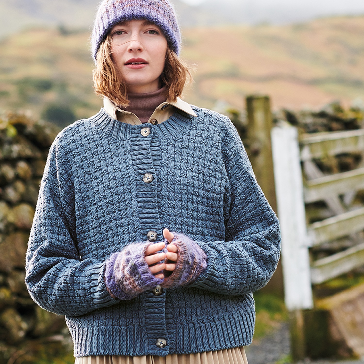 Strickanleitung Jacke aus Essentials Merino dk: Frau trägt blaue Strickjacke und Mütze in ländlicher Umgebung.