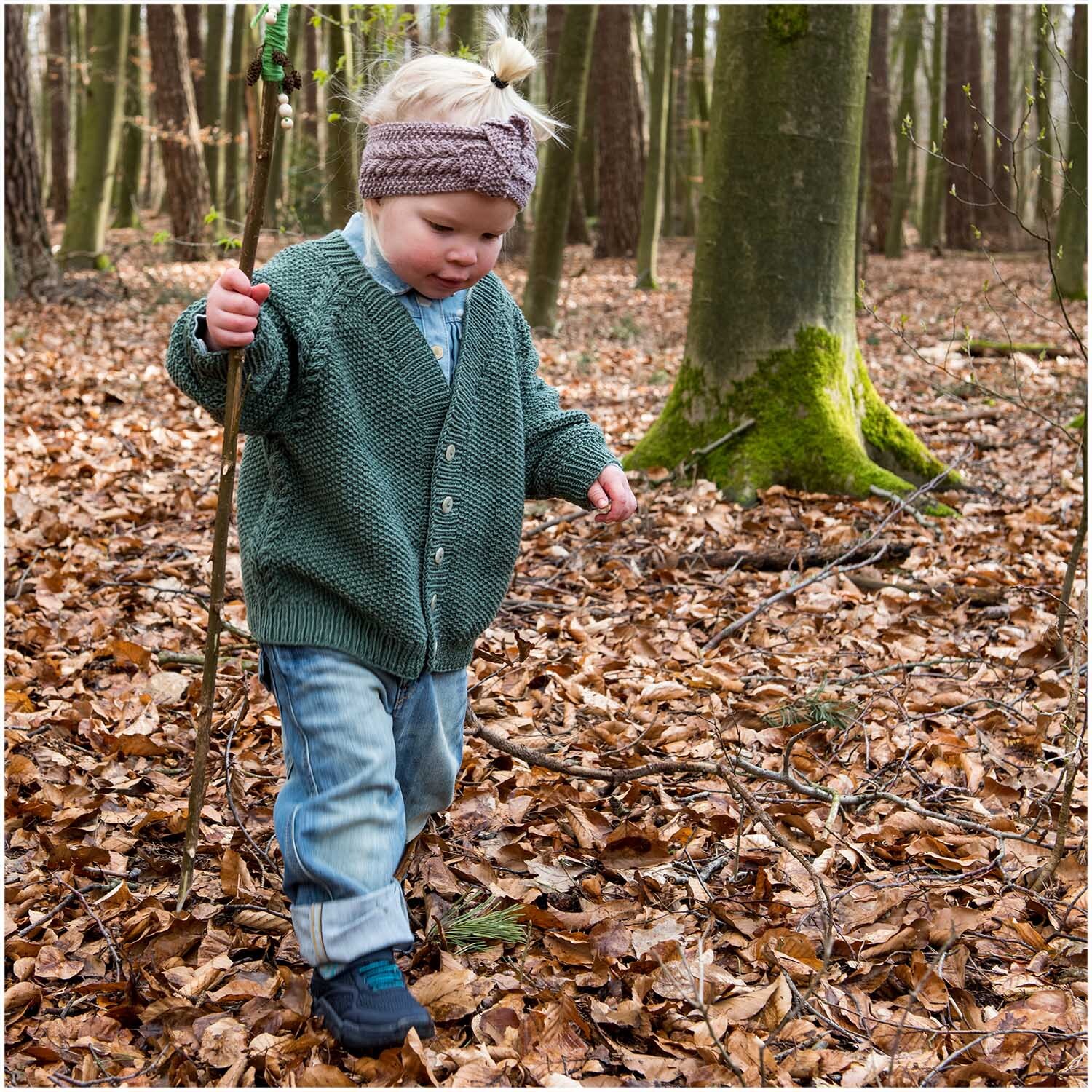 Essentials Merino dk: Kind im Wald mit Strickjacke aus Rico Design Wolle, umgeben von Herbstlaub.