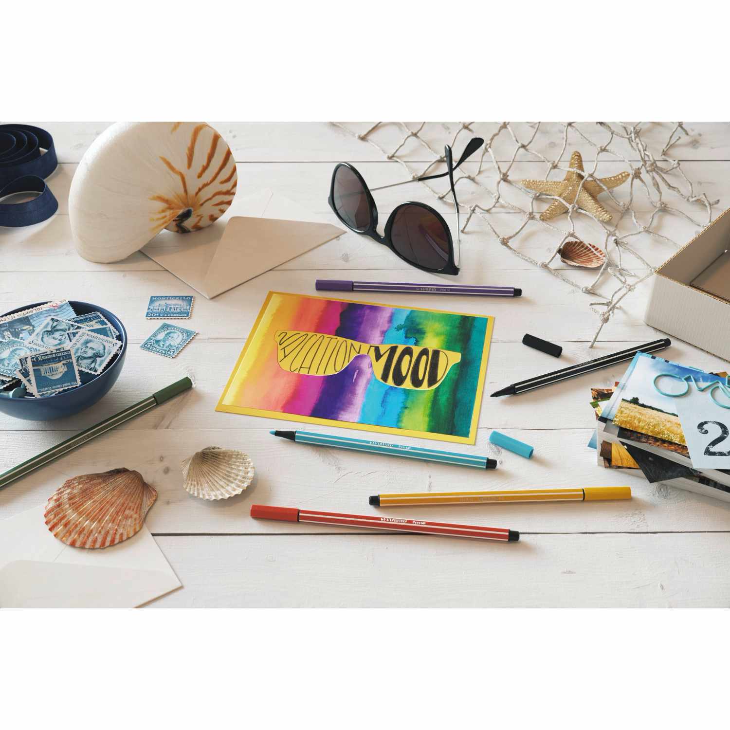 Pen 68: Bunte Stifte und Muscheln auf einem Tisch, mit einer farbenfrohen Zeichnung und Sonnenbrille.