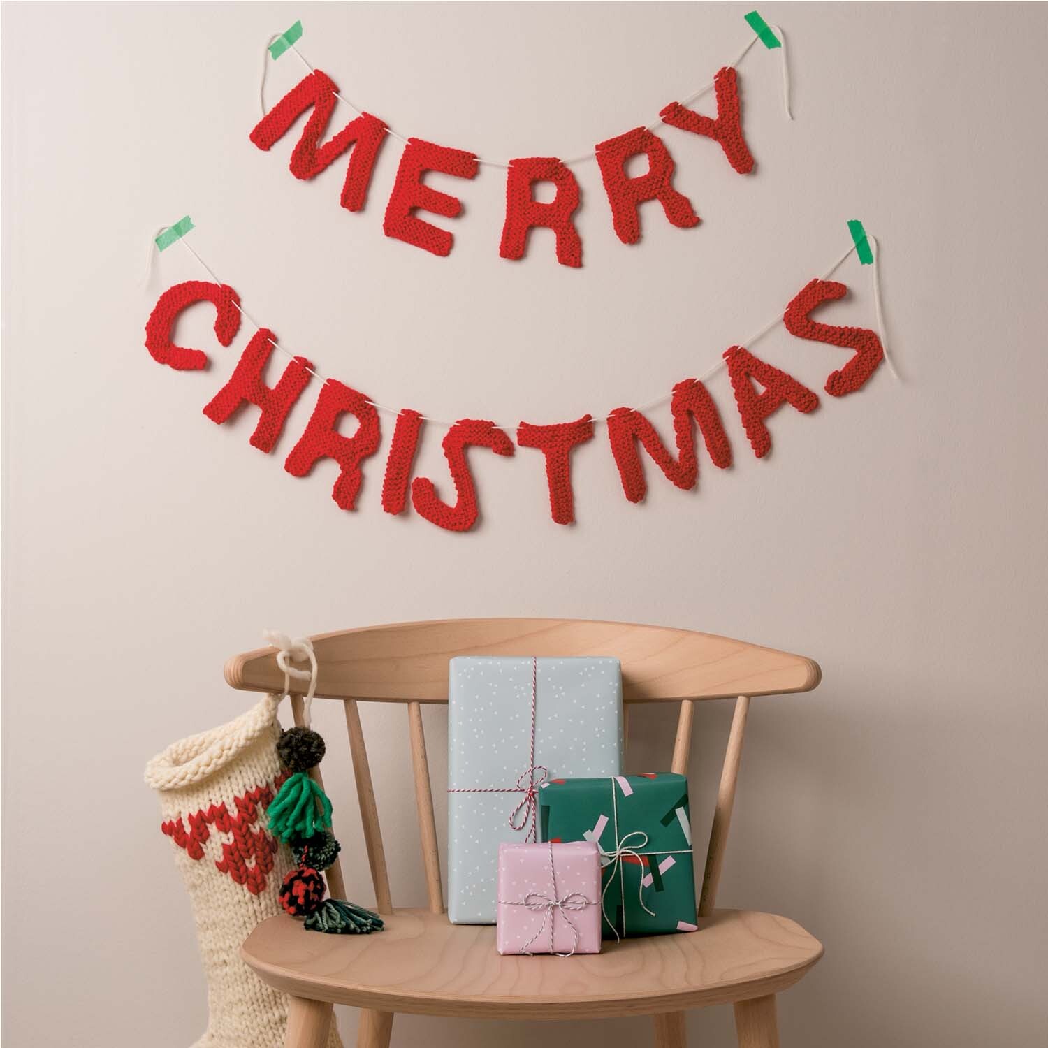 Strickanleitung Merry Christmas Girlande: Rote gestrickte Buchstaben an der Wand, darunter Geschenke auf einem Stuhl.