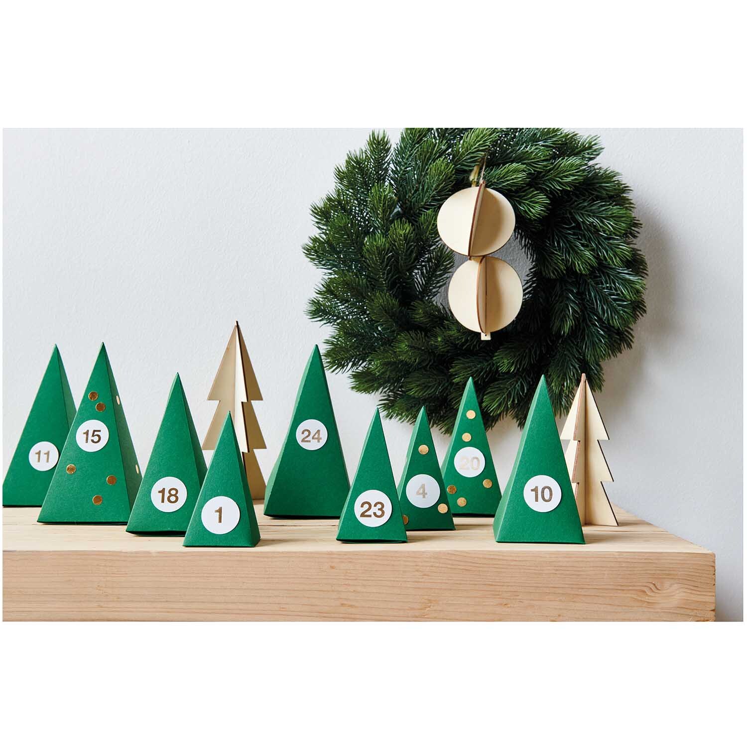 Paper Poetry Adventskalender Bäume 24 Stück: Grüne und holzfarbene Tannen auf Holzbrett, dekoriert mit Zahlen.